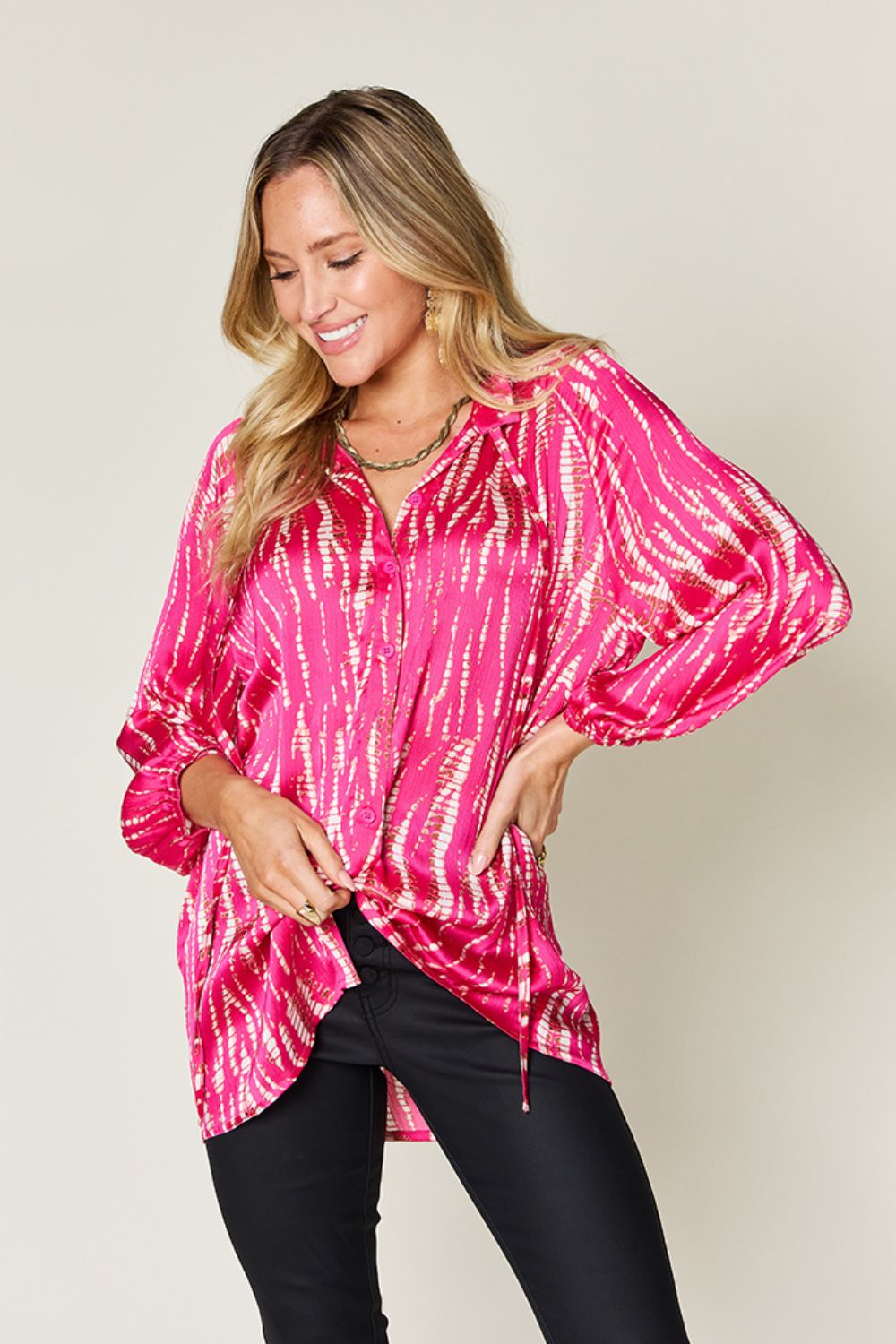 A woman wearing the Double Take Full Size Printed Button Up Long Sleeve Shirt in vibrant pink polyester and black pants stands against a plain background while touching her hair.