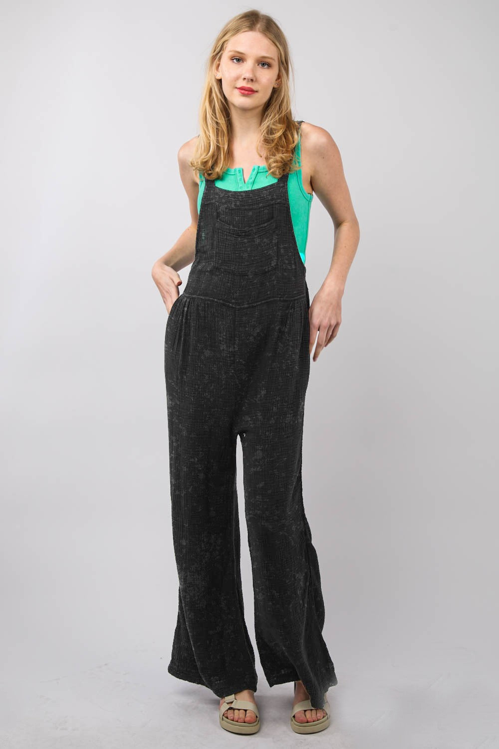 A person wearing the VERY J Texture Washed Wide Leg Overalls and a green top stands against a gray background, paired with sandals, presenting a stylish and relaxed look.