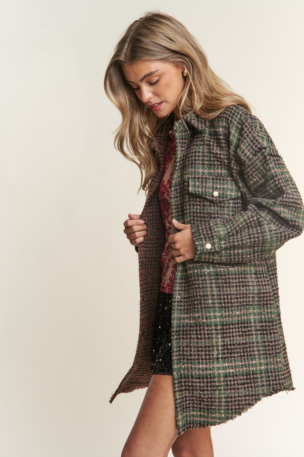 A woman wearing an eclectic mix poses against a plain background, showcasing an ideal fall wardrobe ensemble. She dons a mixed-patterned oversized jacket over a red floral sheer top and black sequined shorts. The J.NNA Snap Down Tweed Plaid Contrast Longline Shacket adds texture and depth to her look.