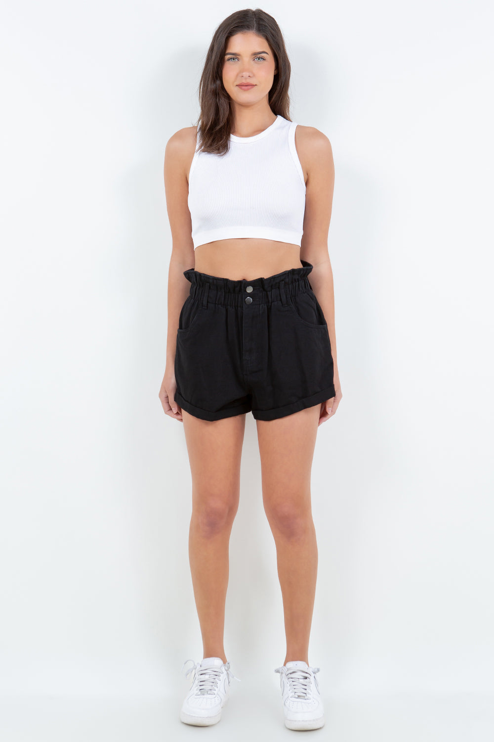 Person wearing black American Bazi High Waist Paper Bag Shorts standing with legs apart. Made from lightweight fabric, these shorts feature large front pockets and a button closure. White sneakers are visible, making this a perfect addition to your summer wardrobe.