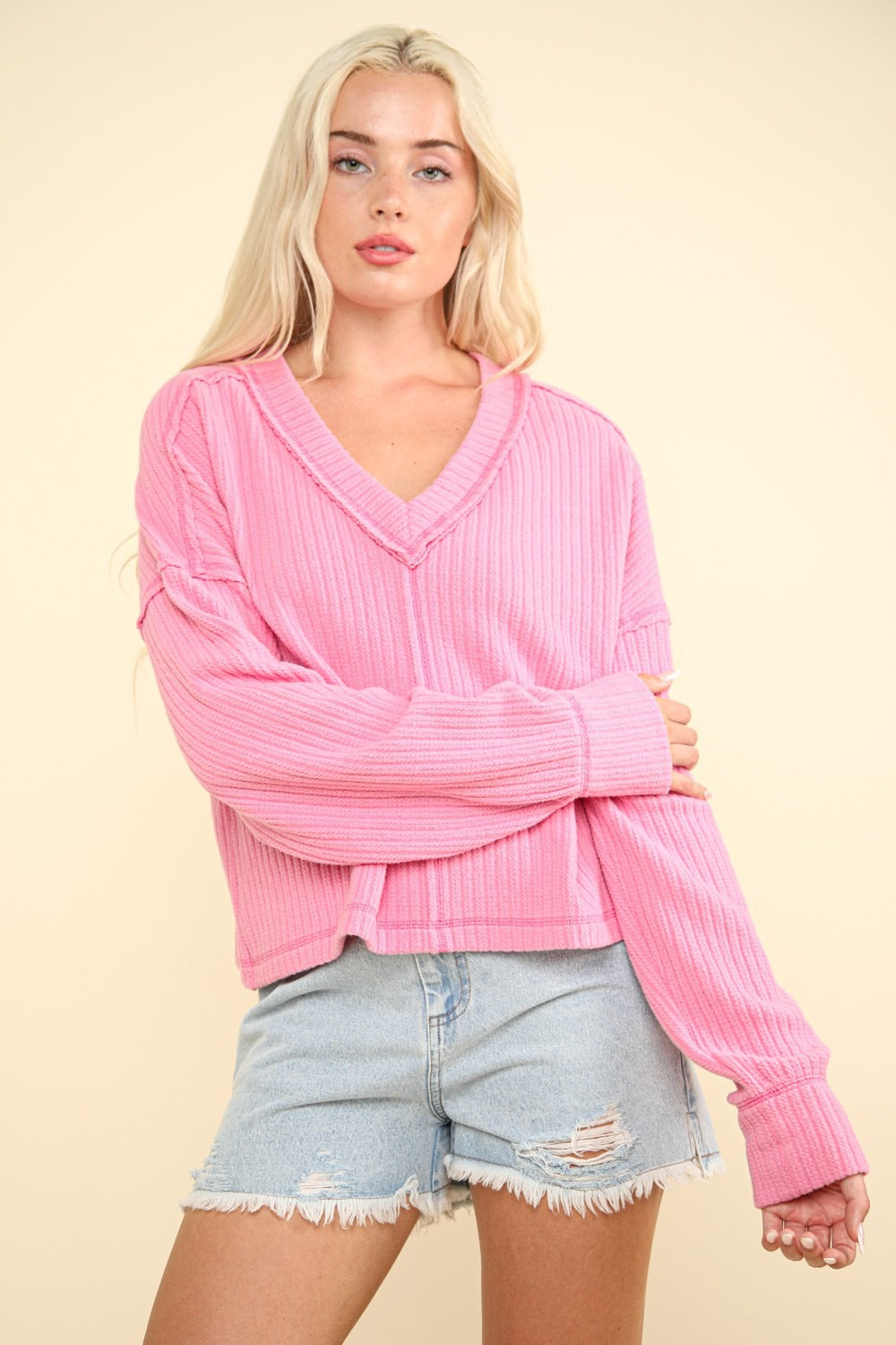 A person with long blonde hair wears a VERY J Exposed Seam V-Neck Ribbed Knit Top in pink and denim shorts, standing against a plain beige background.