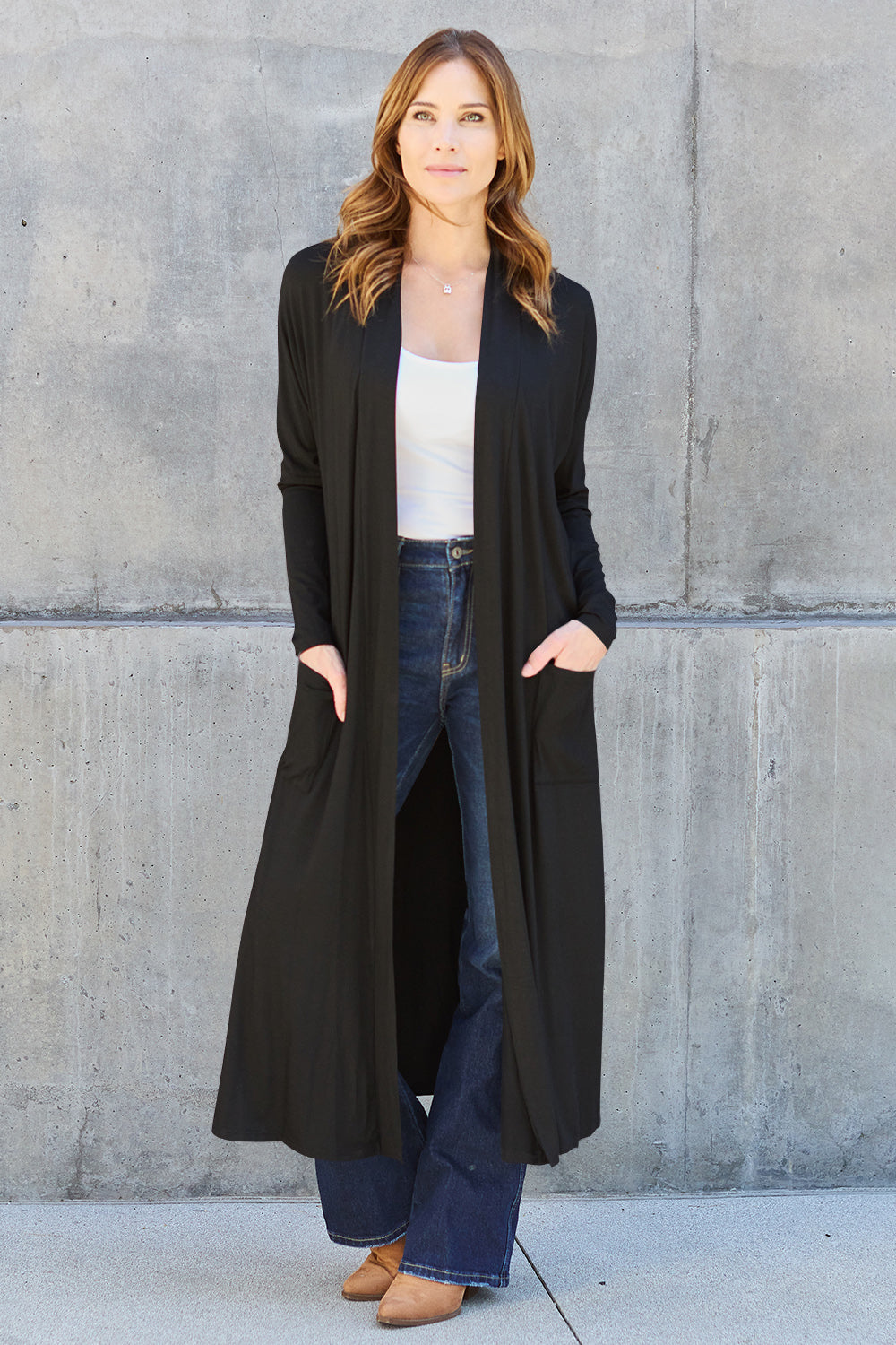 A woman stands against a concrete wall, wearing the Basic Bae Full Size Open Front Long Sleeve Cover Up made from rayon spandex, a white top, blue jeans in her perfect size, and brown shoes with her hands in her pockets. Machine wash cold for best results.