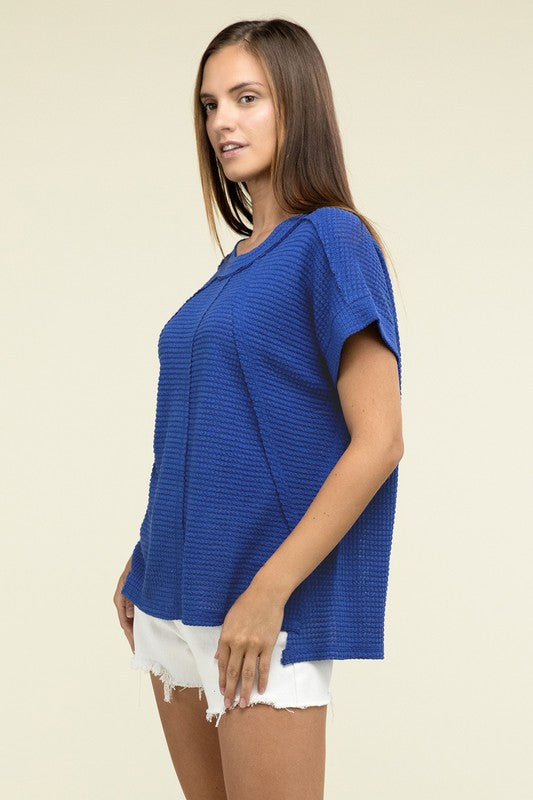 A woman with long brown hair is wearing a purple Brushed Waffle Exposed-Seam Short Sleeve Top and blue jeans, standing against a plain beige background. This casual wardrobe choice, with its short sleeves and side slits, is perfect for everyday wear.