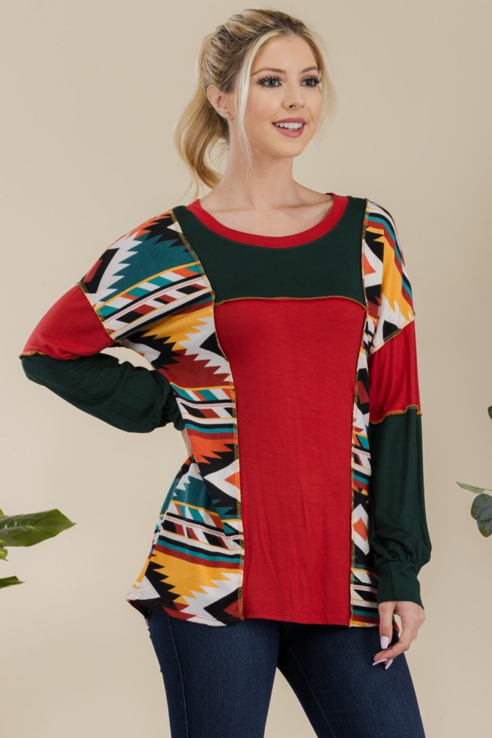 A woman stands against a plain background, wearing the Celeste Full Size Exposed Seam Printed Color Block T-Shirt with vibrant red, green, and yellow geometric sections, paired with dark jeans. She smiles, her hands resting in her jeans' pockets.