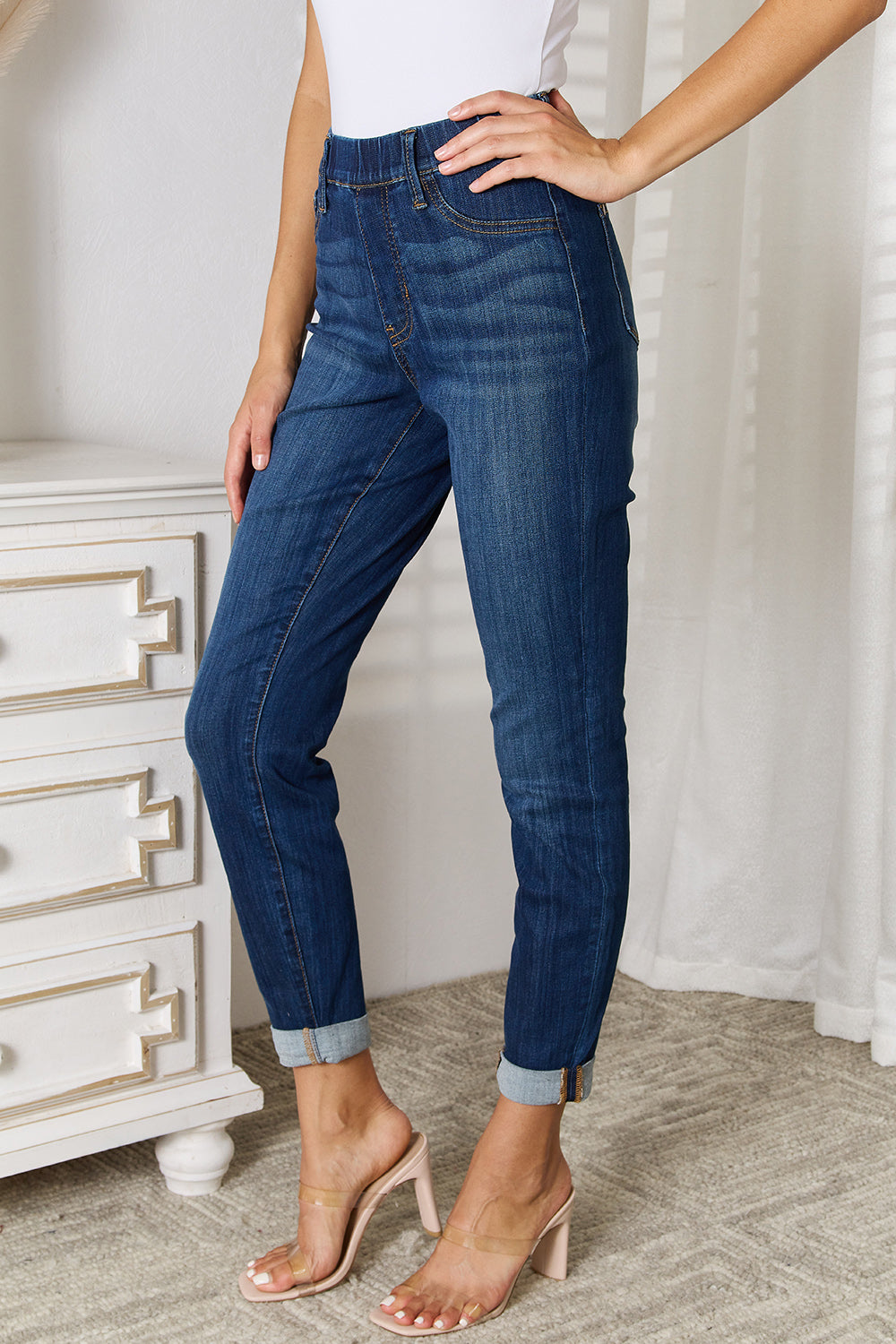 A person wearing Judy Blue Full Size Skinny Cropped Jeans with rolled-up cuffs and clear heels stands near a white dresser adorned with a decorative vase for a modern look.