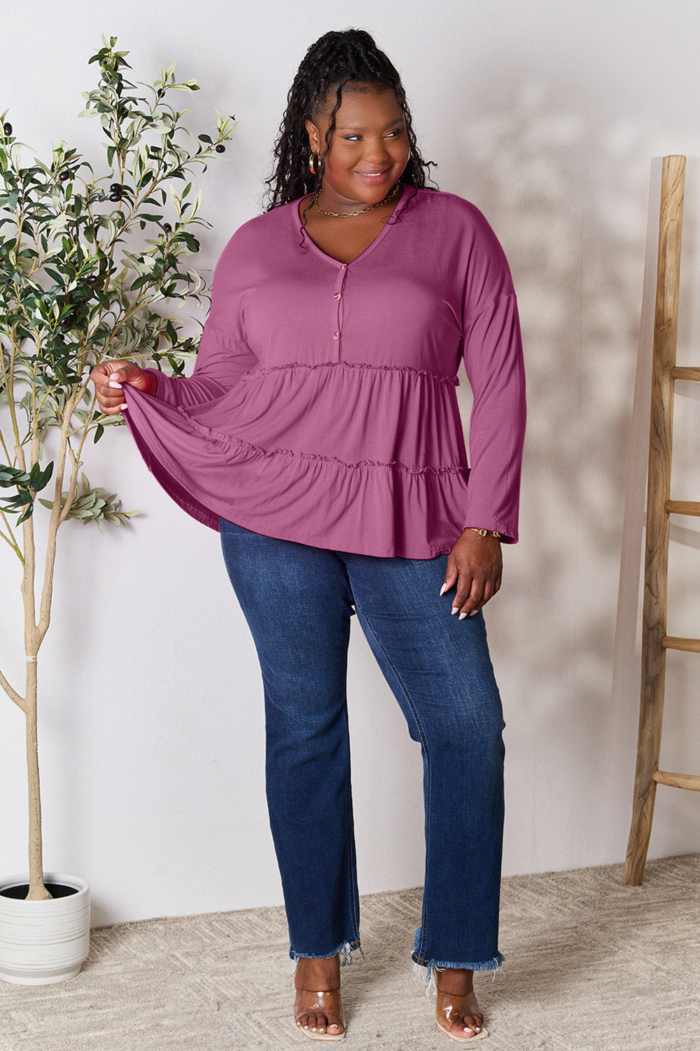 Person stands indoors near a plant, wearing the Double Take Half Button Long Sleeve Ruffle Hem Blouse in gray and blue jeans, smiling while looking to the side. The slightly stretchy fabric of the blouse adds comfort while its buttoned design offers a touch of elegance.