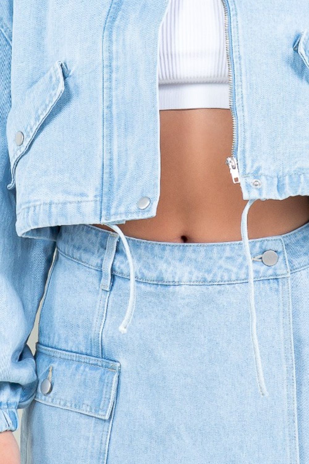 A woman wearing an American Bazi Drawstring Waist Crop Denim Jacket over a white top and a matching light denim skirt stands facing forward against a plain background.