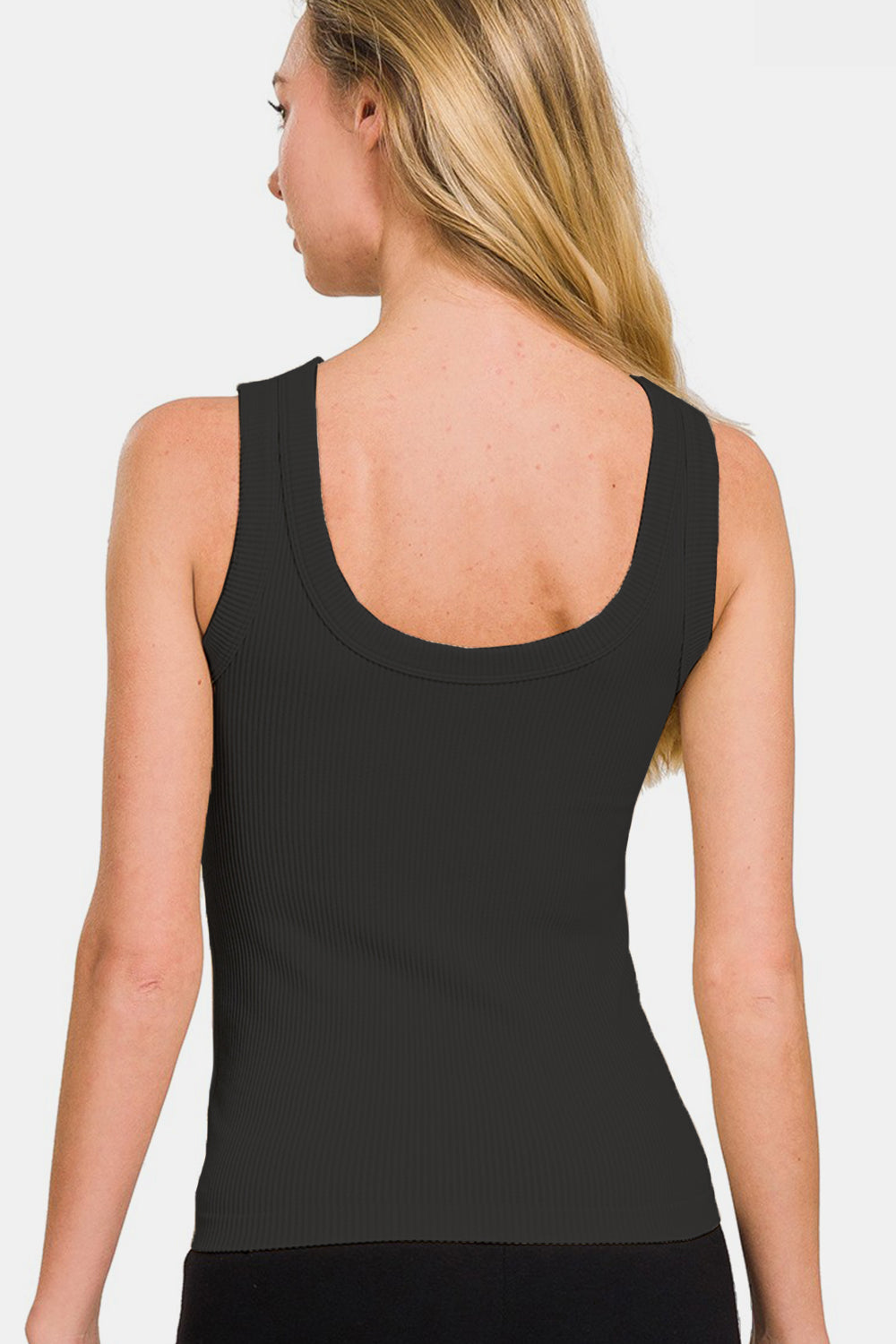 A woman with long blonde hair, adorned in a versatile, black Zenana 2 Way Neckline Washed Ribbed Tank and brown bottoms, stands with one hand on her hip against a plain background.