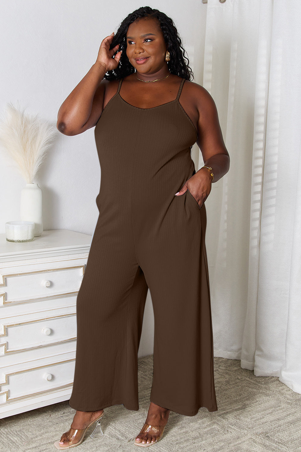 A woman in a stylish Basic Bae Full Size Spaghetti Strap V-Neck Jumpsuit stands indoors near a white dresser adorned with decorative items, smiling with one hand in her pocket.