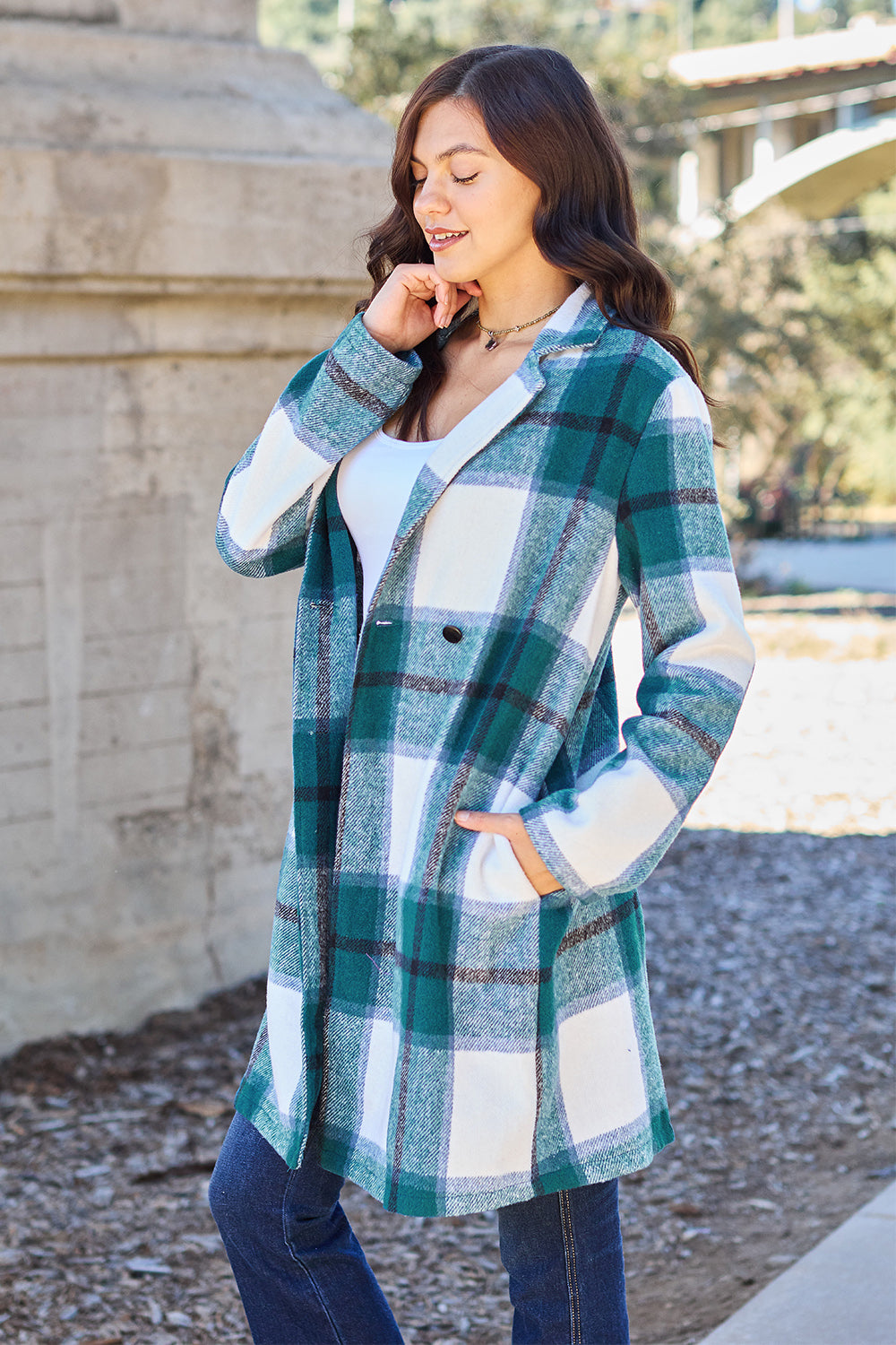 A woman stands on a sidewalk wearing the Double Take Full Size Plaid Button Up Lapel Collar Coat in pink and white over a white top and blue jeans, with her right hand lightly touching her hair. The polyester fabric of the coat makes it durable and perfect for an easy machine wash cold.