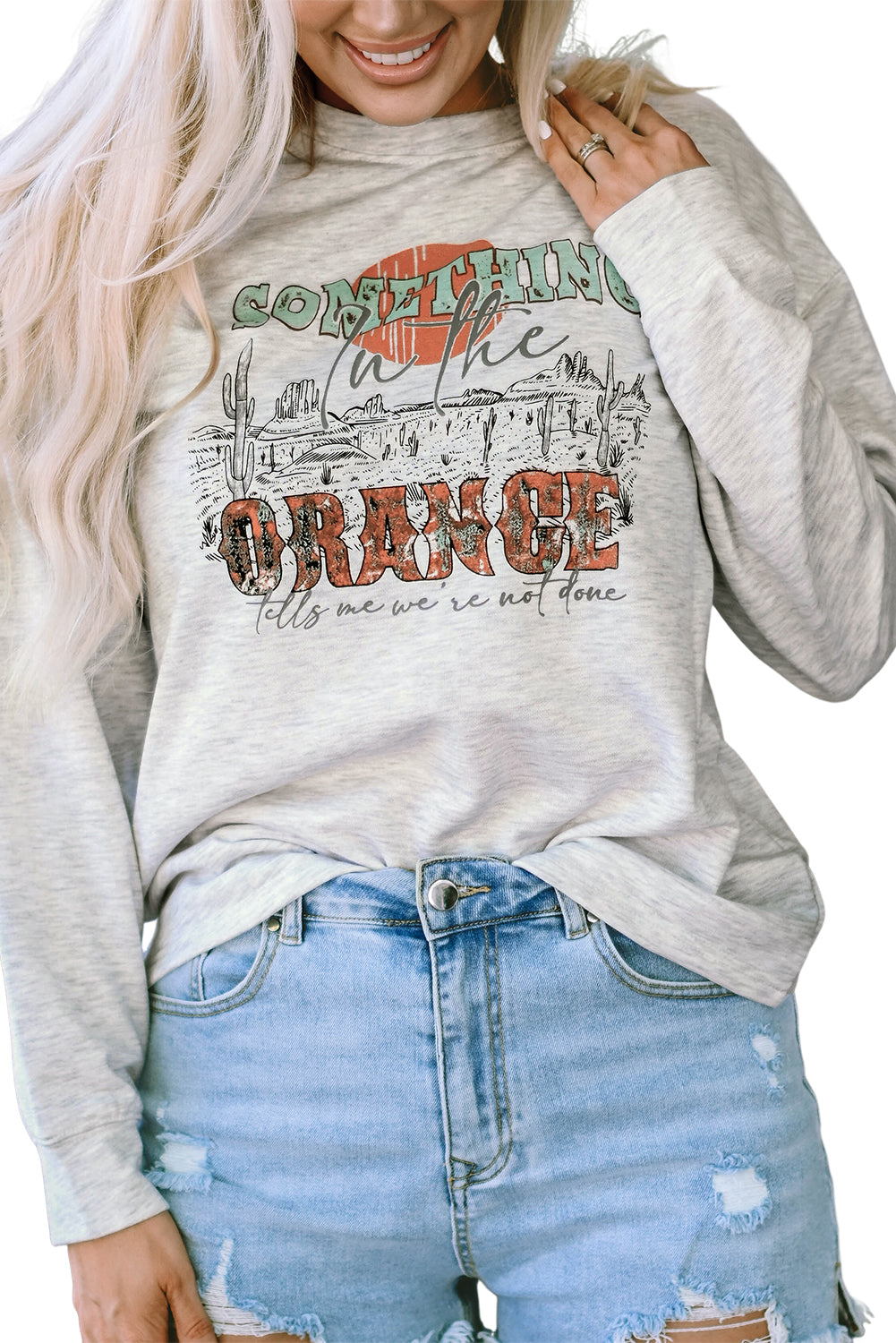 A person with long blonde hair is seen from behind, wearing denim shorts and a Gray SOMETHING ORANGE Graphic Relaxed Sweatshirt featuring a desert sunrise print.