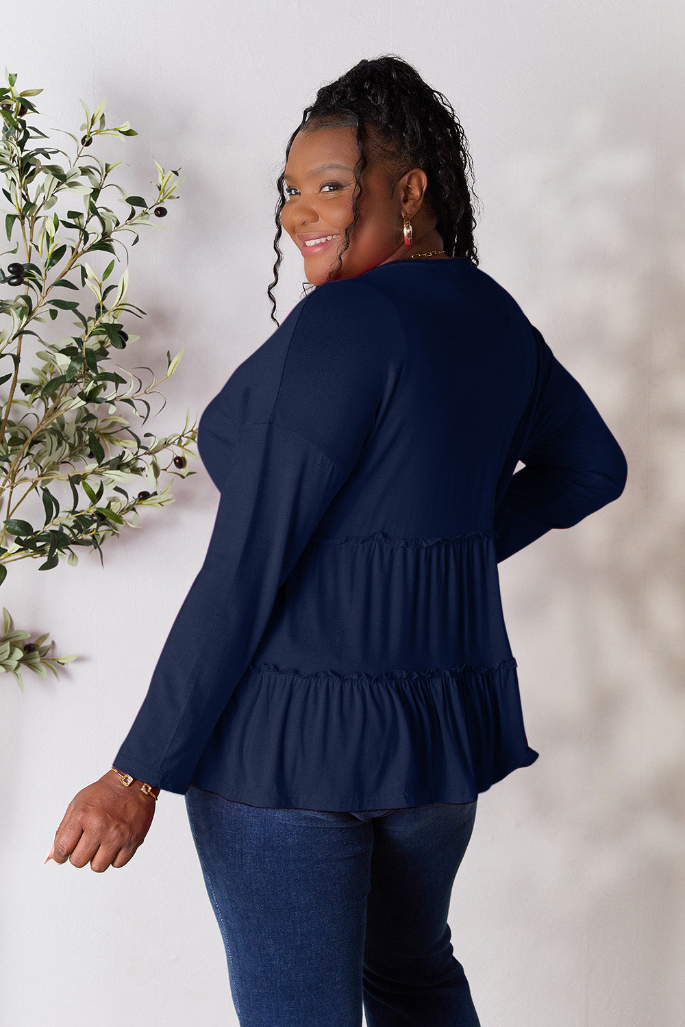 Person stands indoors near a plant, wearing the Double Take Half Button Long Sleeve Ruffle Hem Blouse in gray and blue jeans, smiling while looking to the side. The slightly stretchy fabric of the blouse adds comfort while its buttoned design offers a touch of elegance.