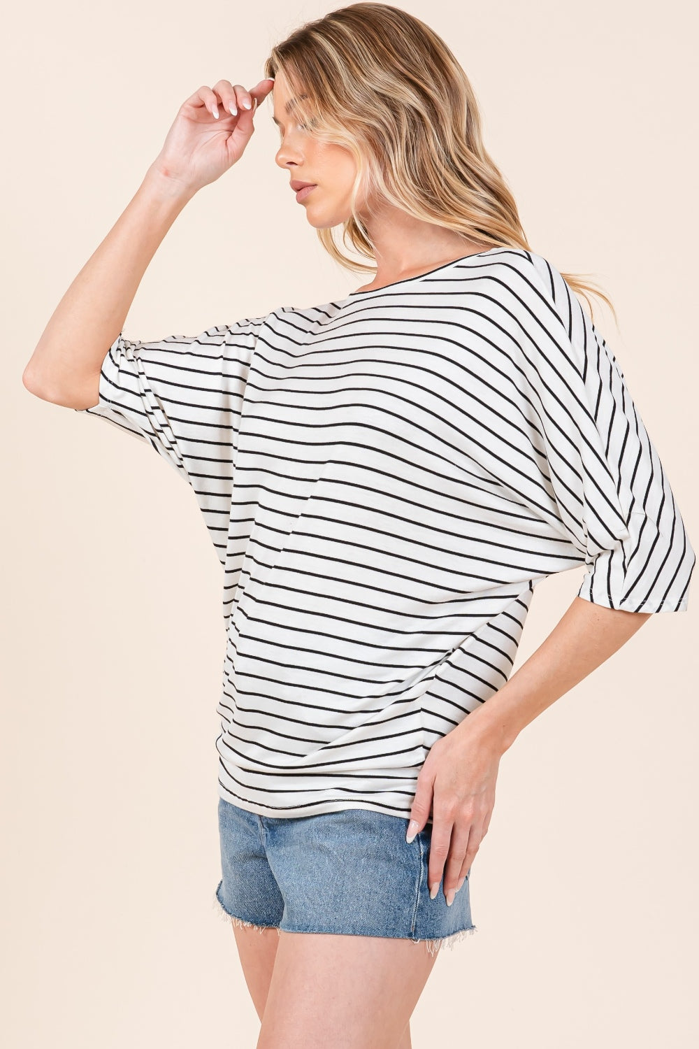 A person is wearing the BOMBOM Striped Boat Neck Dolman Sleeve Top paired with denim shorts, effortlessly embodying a wardrobe staple. The person has their right hand touching their hair and is looking down, against a plain, light-colored background.