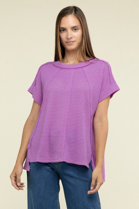 A woman with long brown hair is wearing a purple Brushed Waffle Exposed-Seam Short Sleeve Top and blue jeans, standing against a plain beige background. This casual wardrobe choice, with its short sleeves and side slits, is perfect for everyday wear.