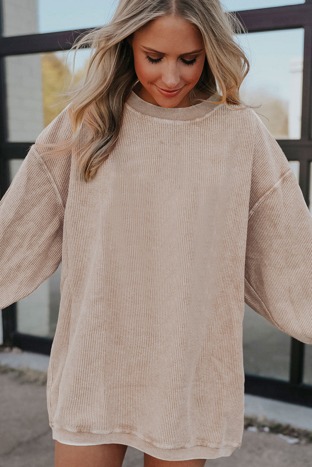 A woman stands outdoors, looking down while touching her hair with one hand. She is wearing an Apricot Ribbed Corduroy Oversized Sweatshirt and white pants, exuding an effortlessly chic and fashionable look.