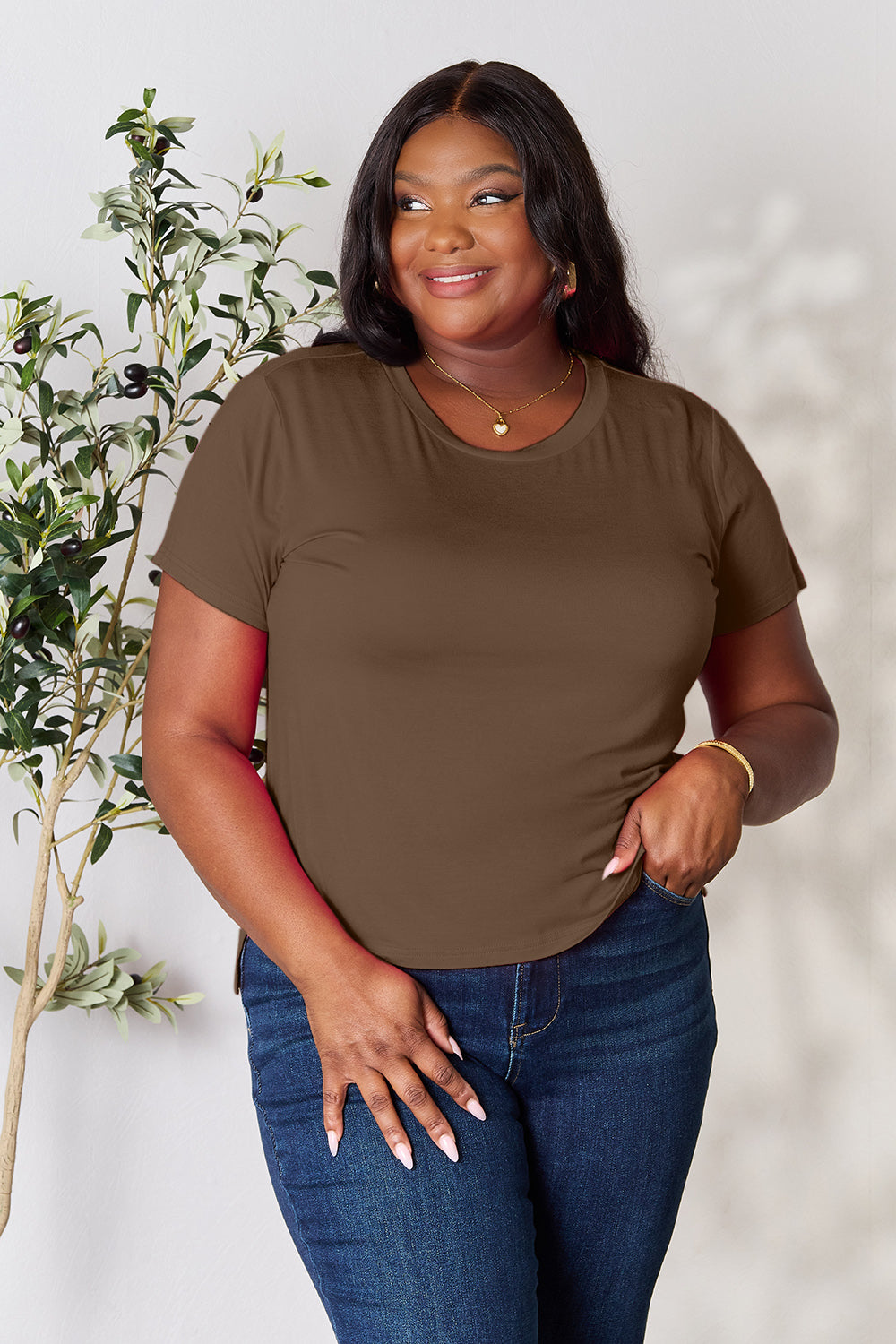 A woman with long hair, smiling with her right hand in her pocket and standing next to a plant, is wearing the Basic Bae Full Size Round Neck Short Sleeve T-Shirt in black along with beige pants made of rayon spandex.