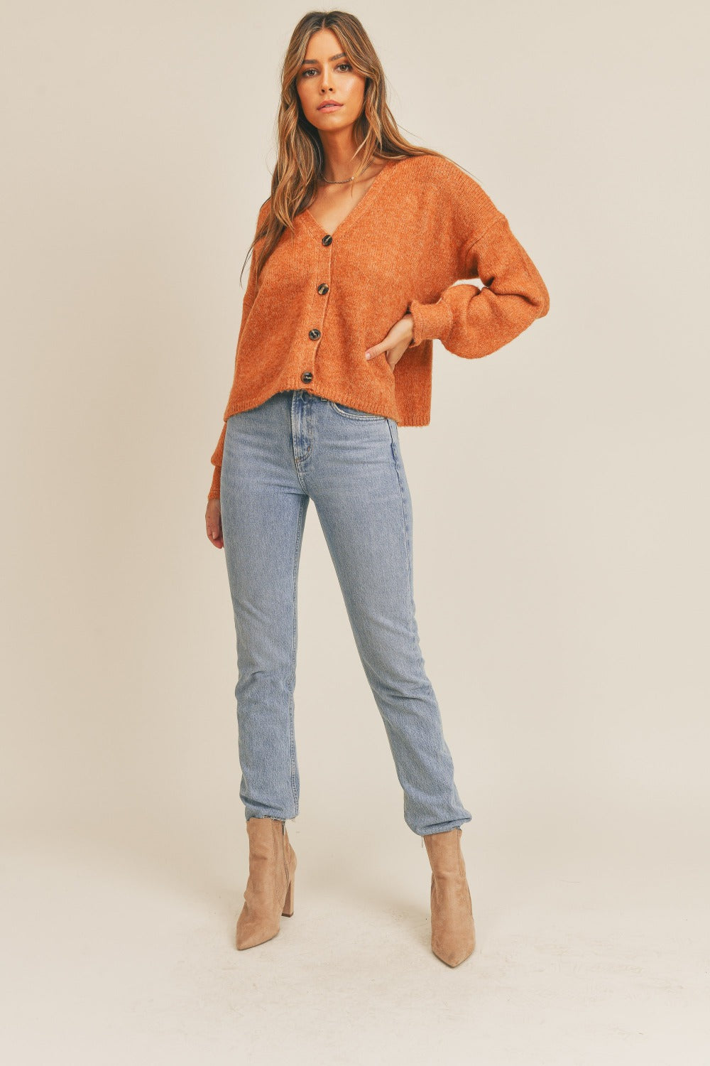 A woman wearing a MABLE Long Sleeve Button Down Sweater Cardigan in burnt orange and light blue jeans looks to her left against a plain background, exuding classic elegance.