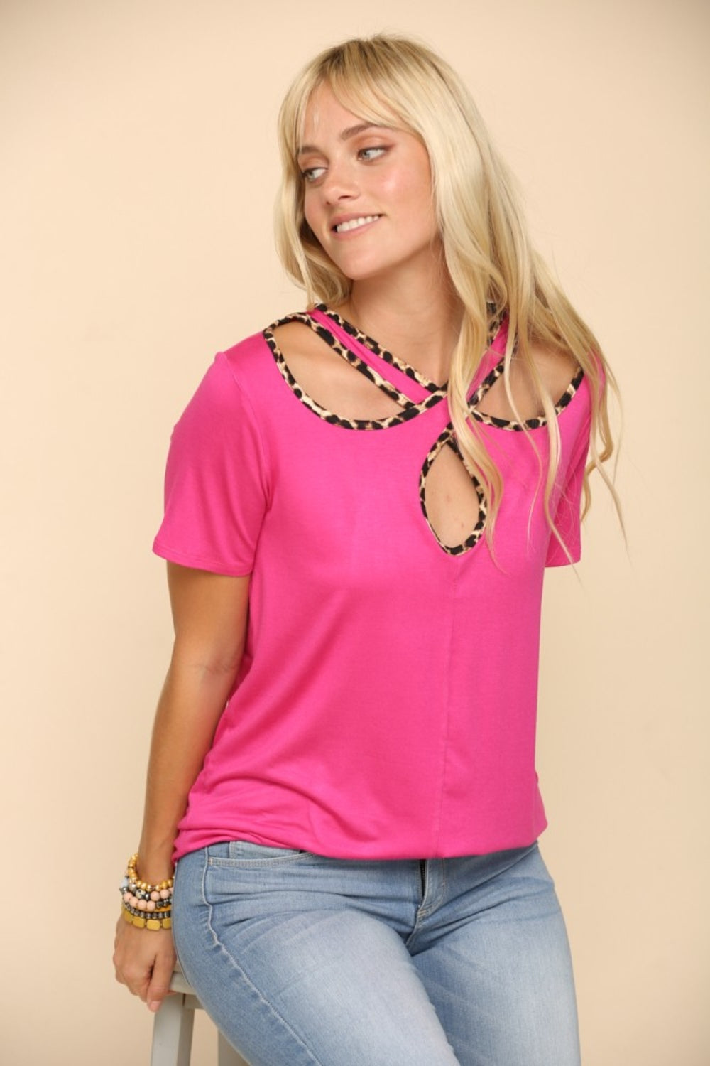 A person showcases the Celeste Full Size Crisscross Cutout Leopard Contrast Trim T-Shirt in bright pink, paired with light blue jeans, posing confidently against a beige background.