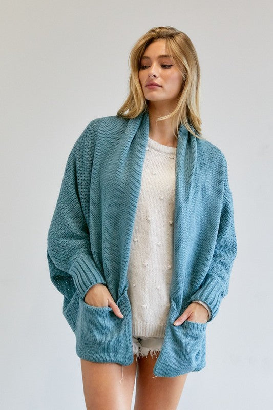 A person stands against a plain background, wearing a Pattern Knit Dolman Sleeve Solid Slouch Cardigan in textured blue over a white sweater with distressed white shorts, hands in cardigan pockets.