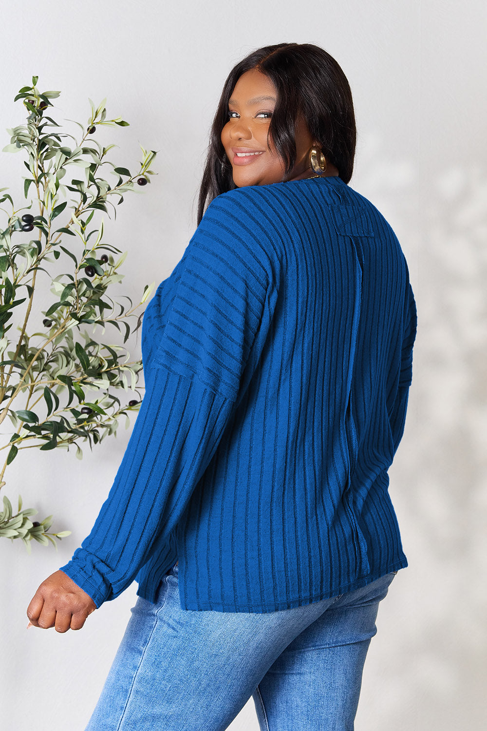 A person wearing the Basic Bae Full Size Ribbed Round Neck Slit Knit Top in blue and beige pants stands and smiles in front of a leafy plant.