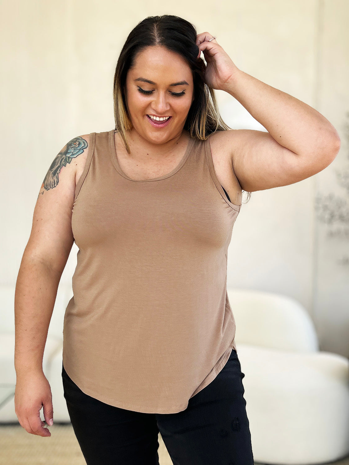 A woman wearing sunglasses, blue jeans, and the Basic Bae Full Size Round Neck Curved Hem Tank is standing in front of a concrete wall, holding her glasses with one hand and carrying a beige shoulder bag. Her basic style complements the slightly stretchy fabric of her outfit, ensuring comfort while maintaining chic simplicity.