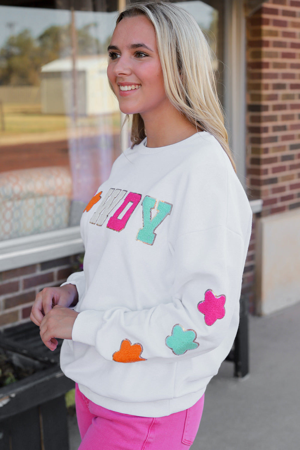 A person poses in the White Howdy Glitter Chenille Patch Graphic Casual Sweatshirt, adorned with colorful star patterns and the letters "WD." She wears light denim shorts and has shoulder-length blonde hair.