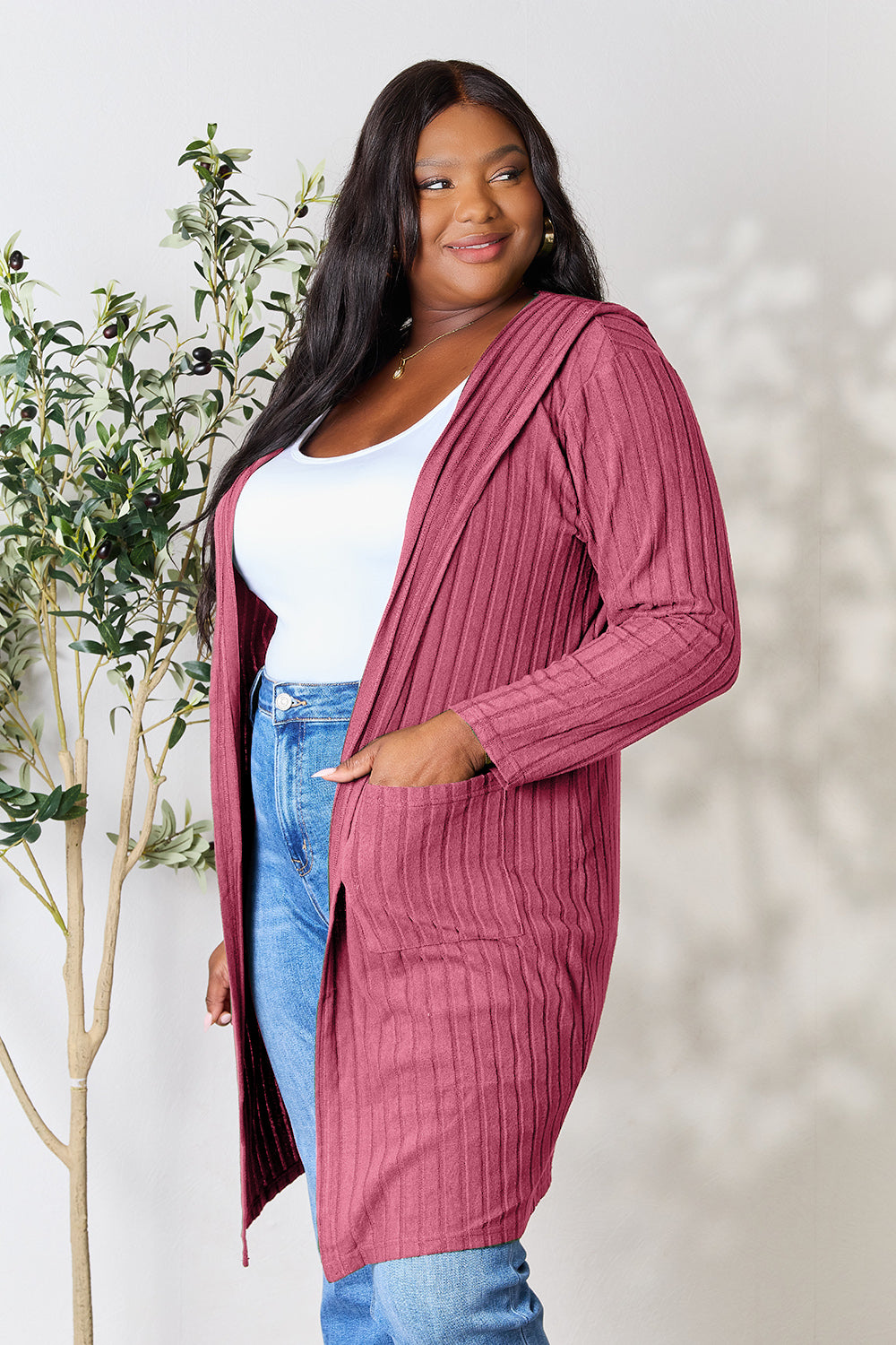 A woman wearing the Basic Bae Full Size Hooded Sweater Cardigan in black, paired with a white top and khaki pants, stands in front of a light background accented by a leafy plant on her left. With one hand tucked into her pocket, she gazes to her right.