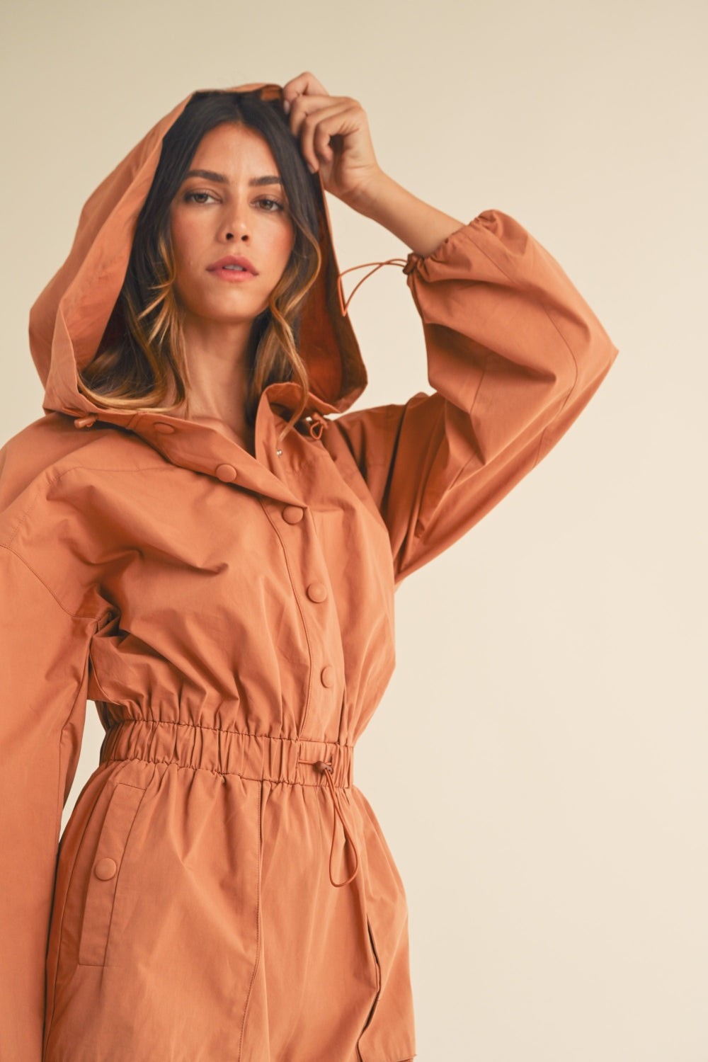 A person wearing the Mable Snap Down Long Sleeve Hooded Romper, featuring a brown color, long sleeves, a snap-down front, and an elastic waistband, is standing against a plain background.