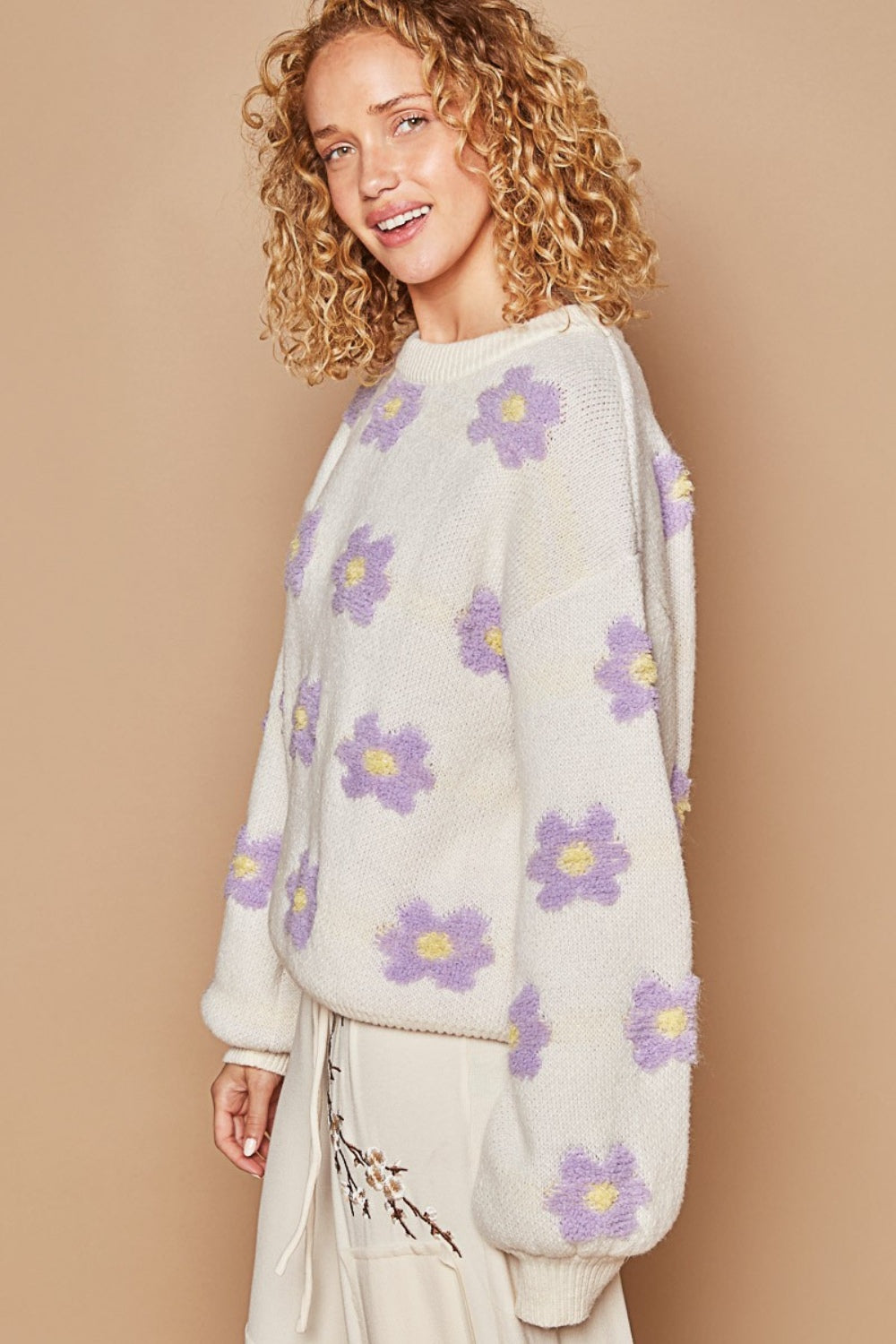 A person with curly hair is smiling against a beige background, wearing an oversized white POL Daisy Pattern Drop Shoulder Sweater featuring a daisy pattern in purple and yellow.