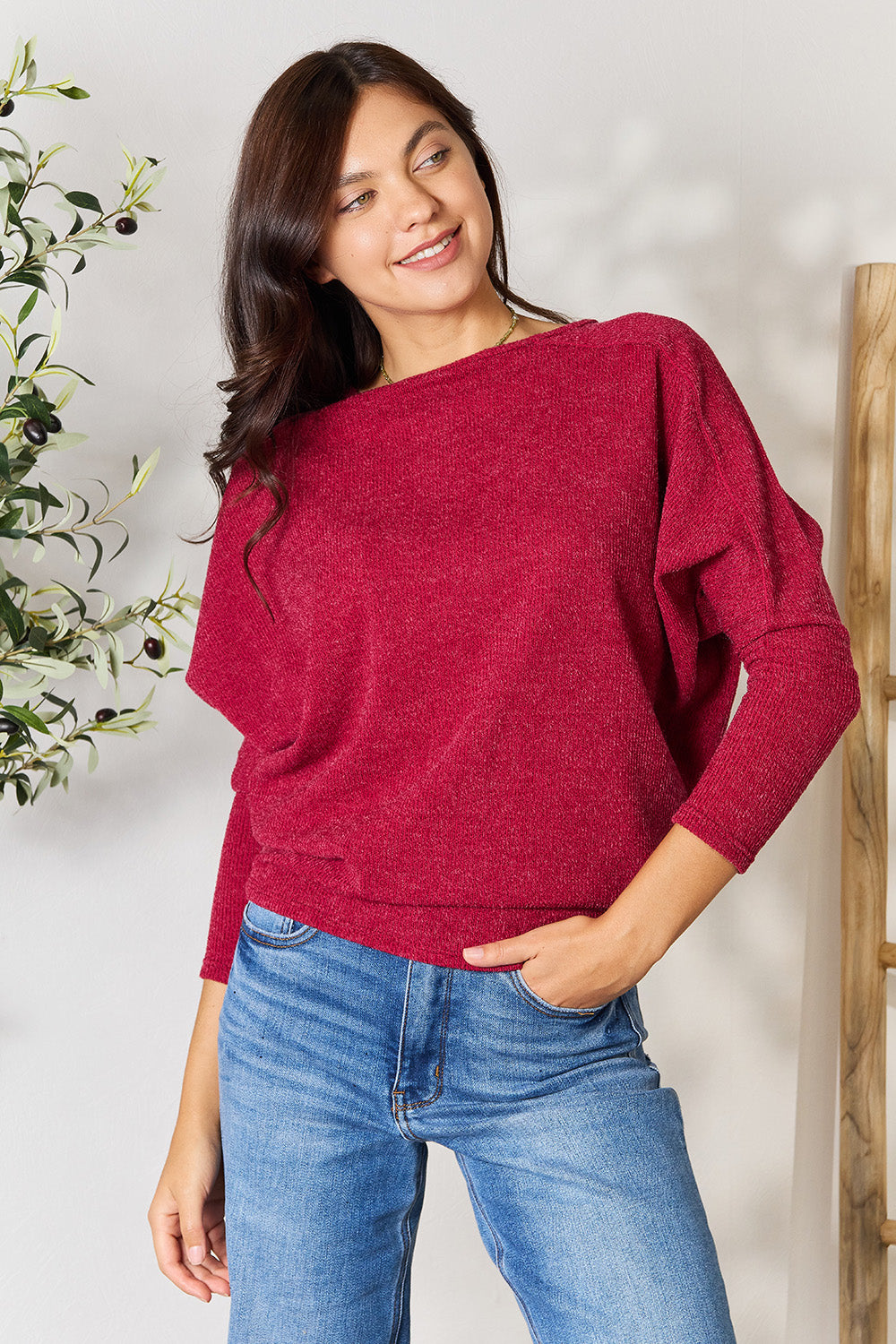 Person wearing a BOMBOM Drop Shoulder Long Sleeve Blouse in red, paired with blue jeans, standing with one hand in their pocket and the other touching their neck, in front of a white wall with a potted plant on the side.