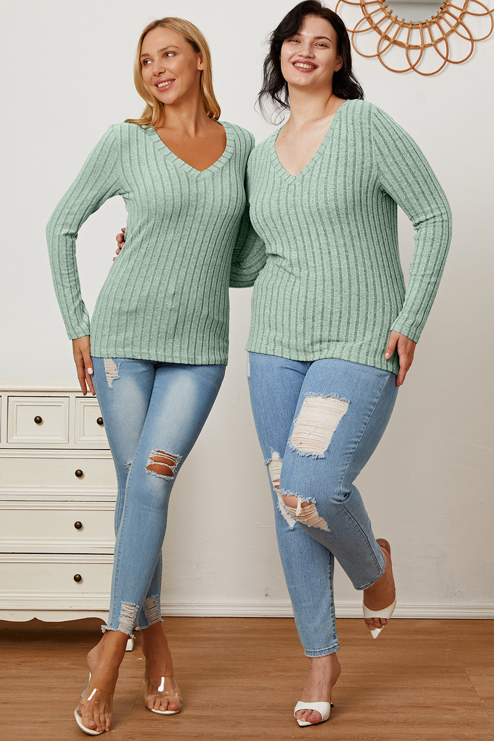 A blonde-haired woman smiles while wearing the Basic Bae Full Size Ribbed V-Neck Long Sleeve T-Shirt, which is light green and slightly stretchy, paired with light blue jeans. She holds one hand near her head and the other near her waist.