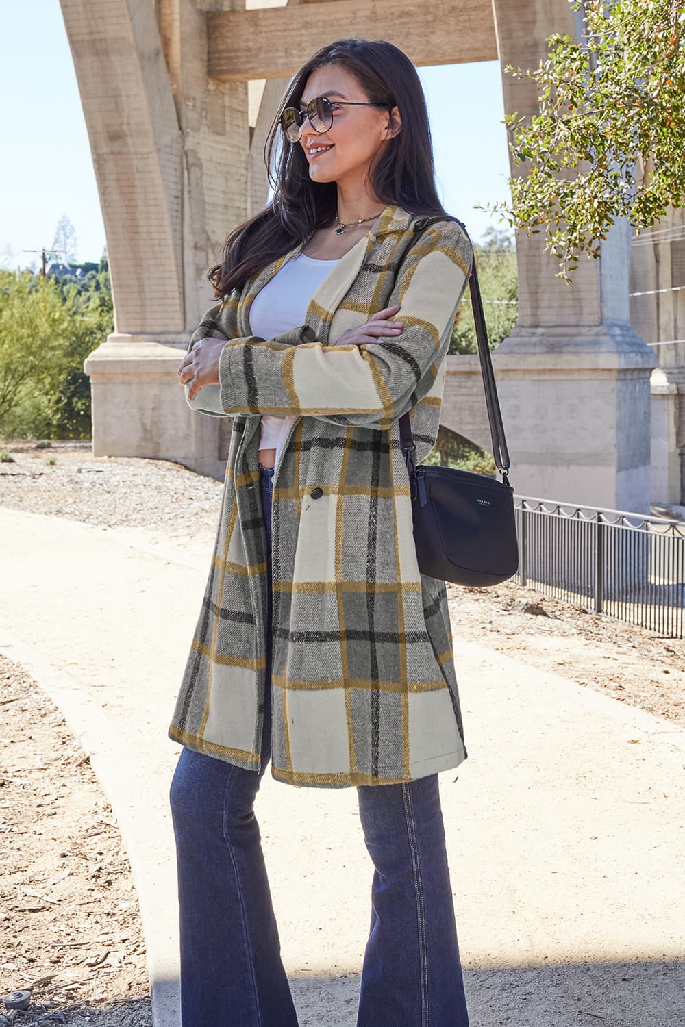 A woman stands on a sidewalk wearing the Double Take Full Size Plaid Button Up Lapel Collar Coat in pink and white over a white top and blue jeans, with her right hand lightly touching her hair. The polyester fabric of the coat makes it durable and perfect for an easy machine wash cold.