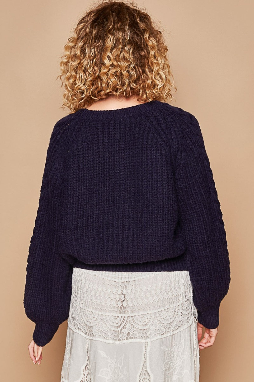 A person is wearing the POL Cable Knit Cutout Long Sleeve Sweater in navy, paired with a white crochet skirt, and is standing against a beige background.