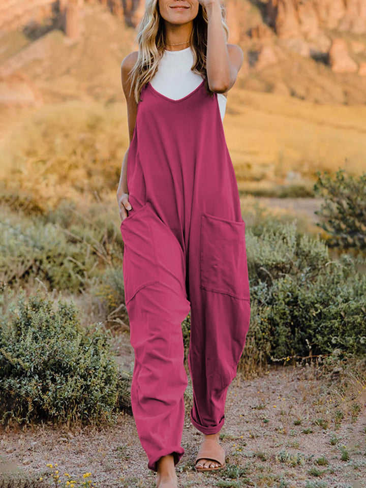 A woman stands outdoors wearing the Double Take Full Size Sleeveless V-Neck Pocketed Jumpsuit in a loose-fitting, imported brown design over a white top. Her surroundings include grass, bushes, and a rocky landscape in the background. The jumpsuit's polyester fabric ensures comfort and durability with every machine wash.