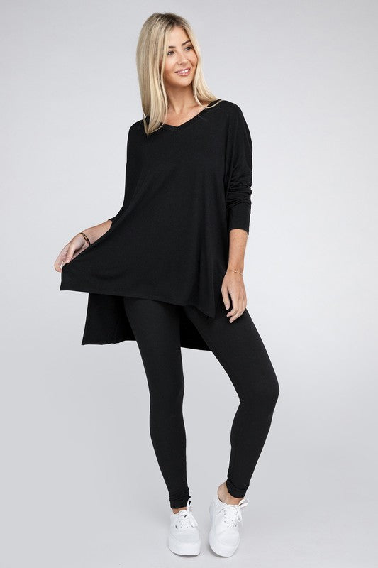 A woman with long, blonde hair is wearing a loose-fitting, brown Dolman Long Sleeve V-Neck Side Slit Hi-Low Hem Top and black pants. She poses with one hand on her hip and a neutral facial expression against a light grey background.