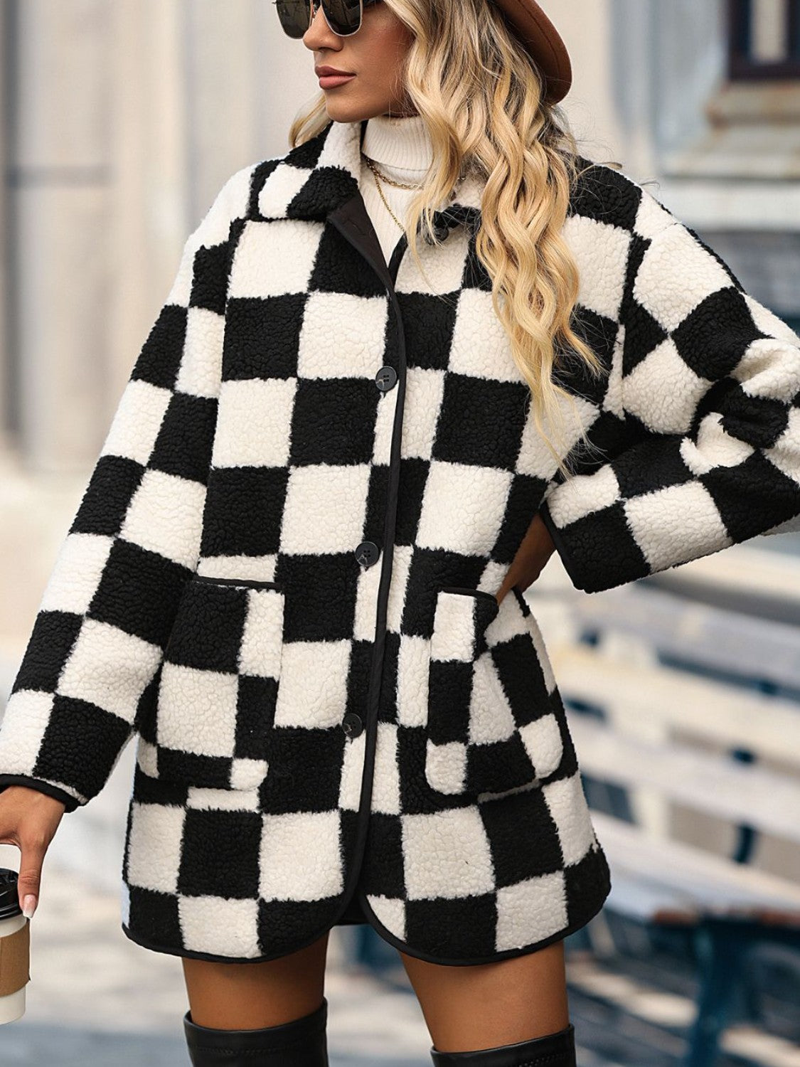 A person wearing the Double Take Full Size Checkered Button Front Coat with Pockets, a white turtleneck sweater, black shorts, and two gold necklaces holds a paper cup. The coat is Basic style and made from 100% polyester.