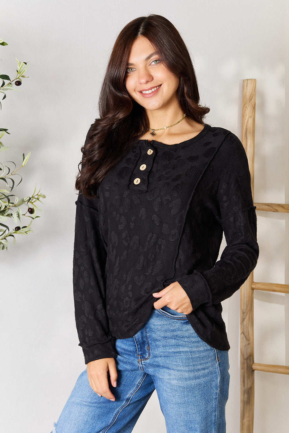 A woman wearing a relaxed fit, black BOMBOM Textured Exposed Seam Buttoned Blouse and jeans stands with one hand in her pocket next to a leafy plant.