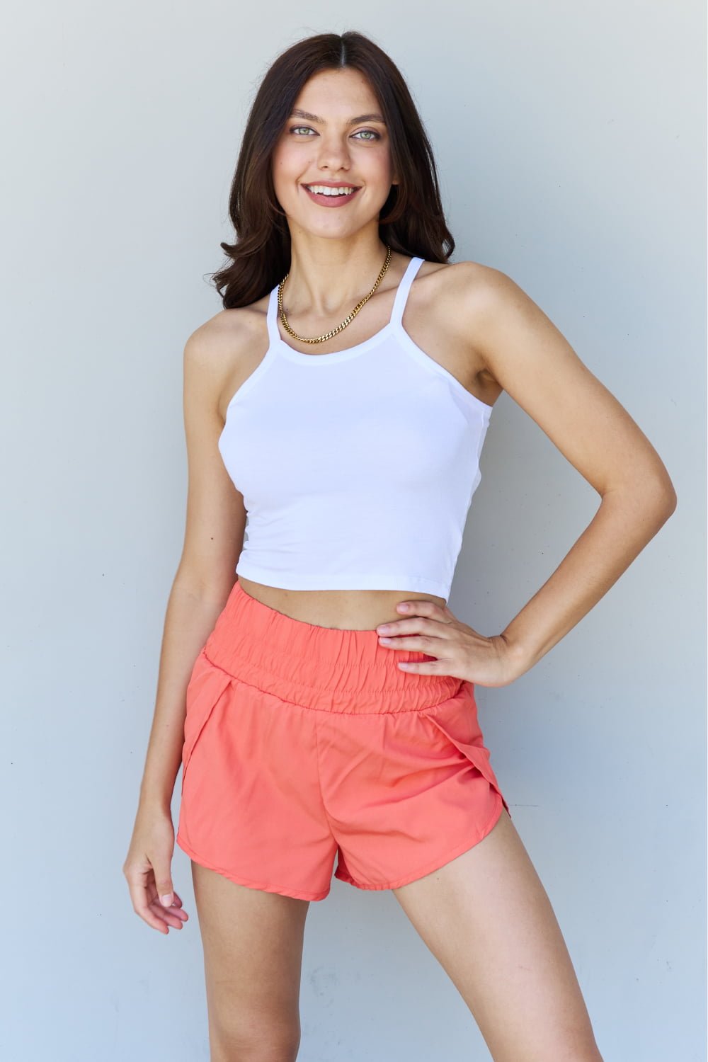 A person wearing the Ninexis Everyday Staple Soft Modal Short Strap Ribbed Tank Top in Off White, paired with coral shorts, stands smiling against a plain backdrop.