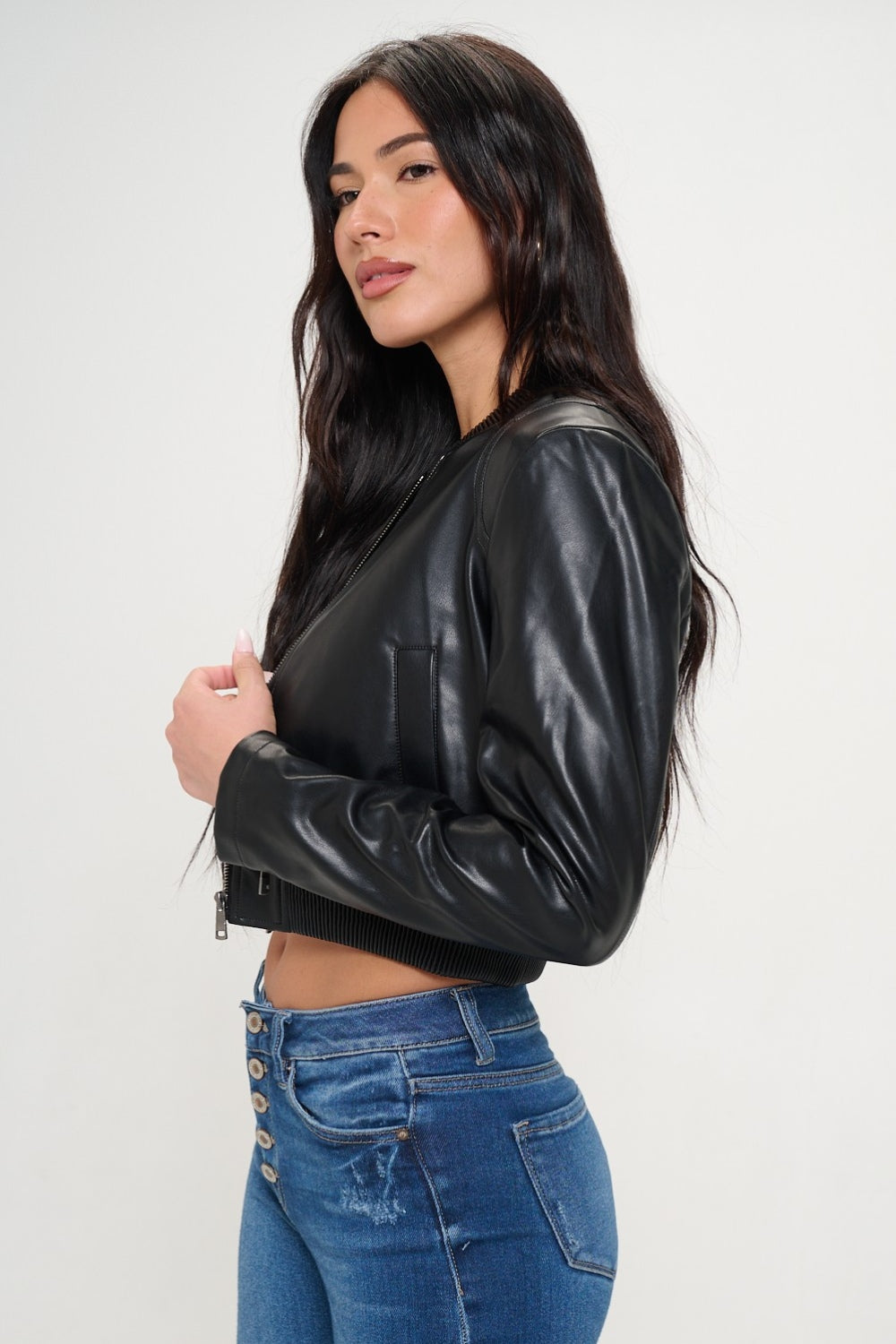 A woman wearing a Coalition LA Zip Up Cropped Bomber Jacket over a black crop top and blue jeans with buttons, looking at the camera with her hands in her jacket pockets.