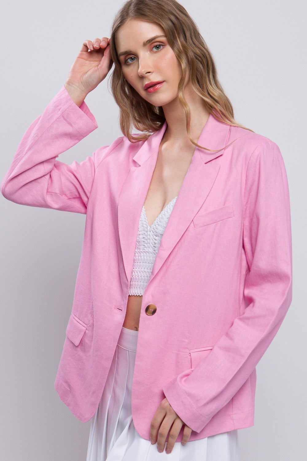 A person with long hair wears a pink Love Tree One-Button Long Sleeve Linen Blazer over a white top, posing with one hand touching their hair.