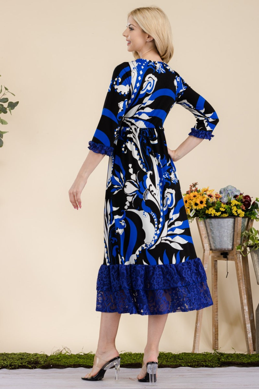 A woman wearing the Celeste Full Size Paisley Print Lace Ruffled Midi Dress stands near a wooden table adorned with a bucket of flowers.