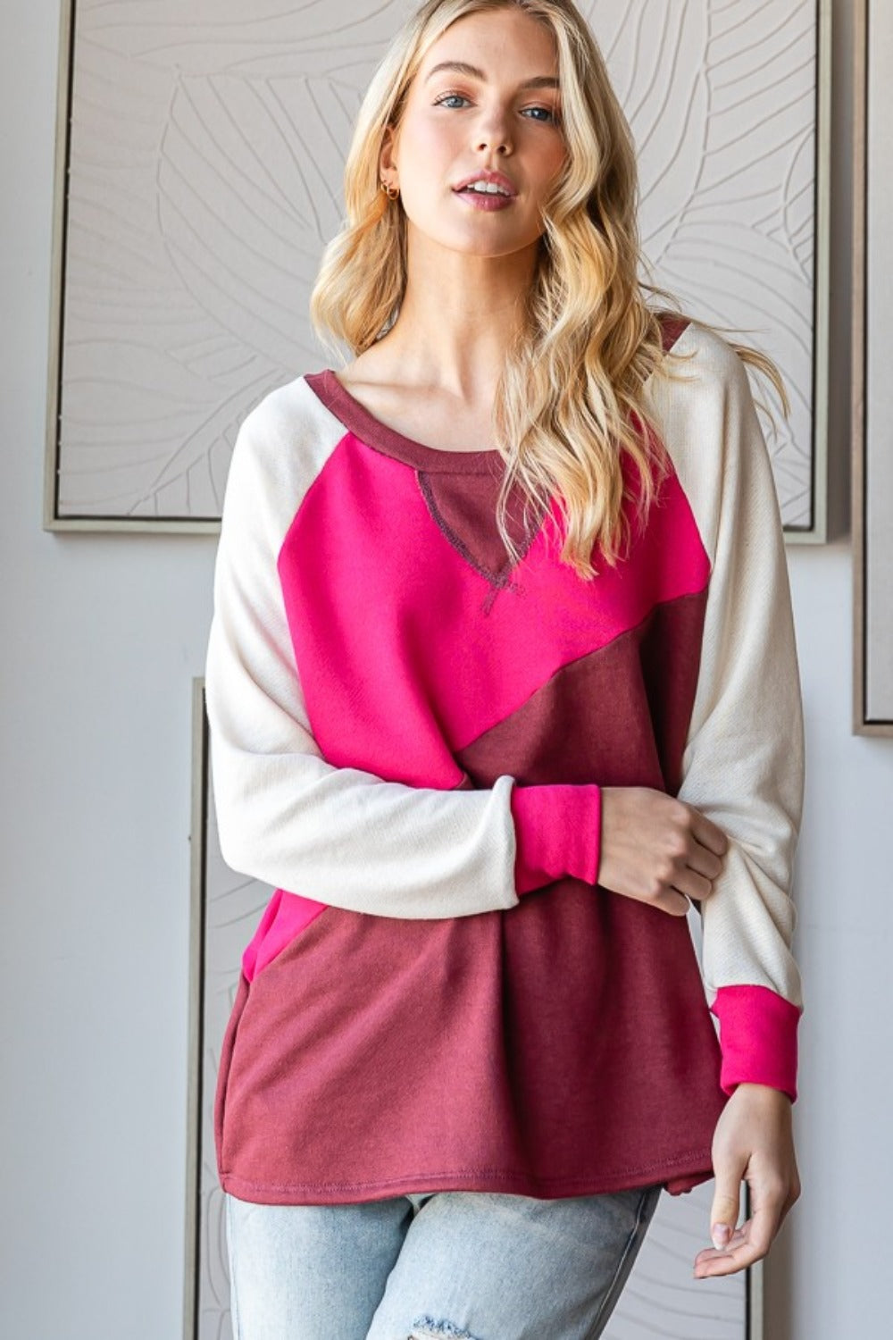 A person with long blonde hair is posing in a HOPELY Color Block Long Sleeve T-Shirt and light blue jeans against a textured wall backdrop.