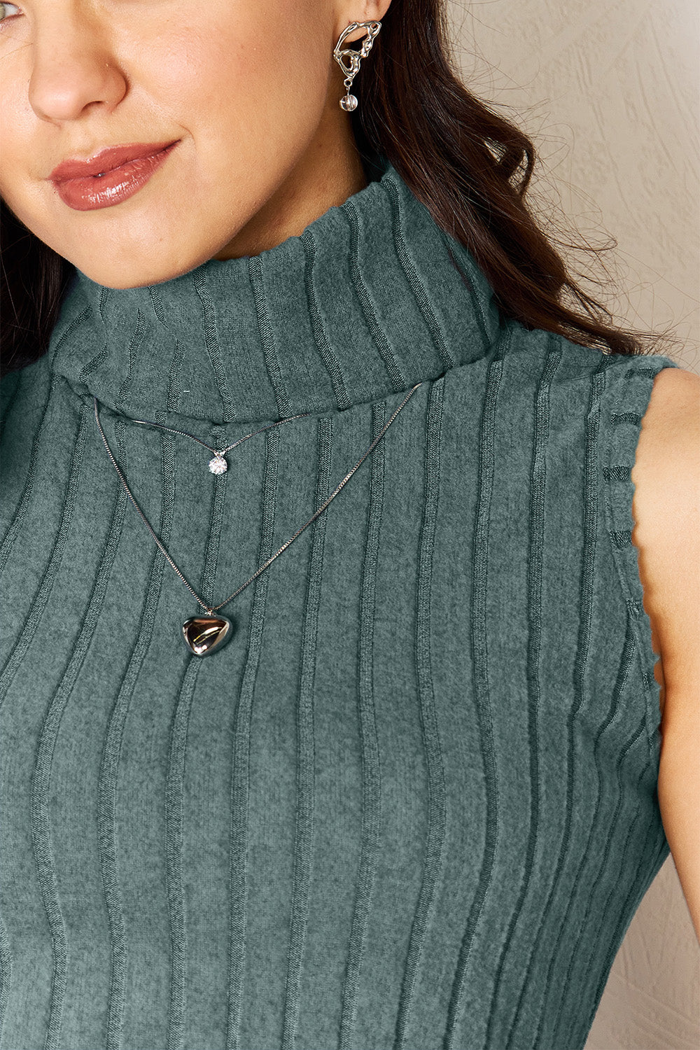 A woman with long brown hair, wearing the Basic Bae Full Size Ribbed Turtleneck Tank in black, along with jeans and necklaces, poses with one hand in her pocket and the other resting on a wooden ladder.