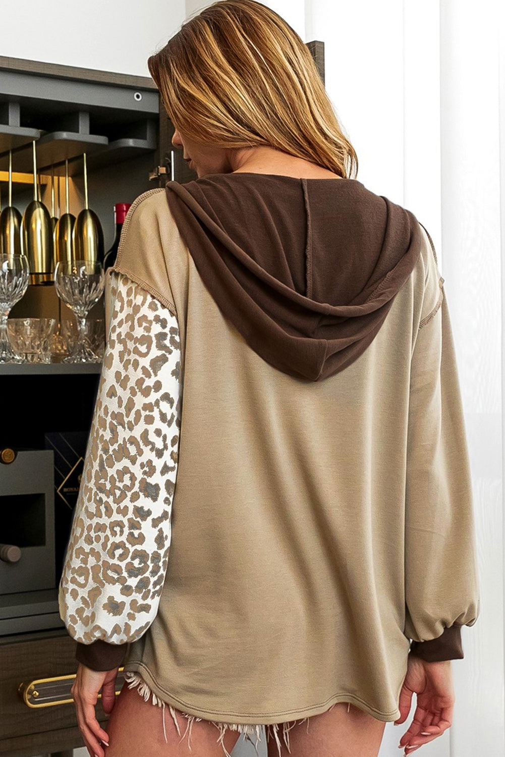 A person with long hair is seen from behind, wearing the Brown Leopard Print Colorblock Chest Pocket Henley Hoodie, which features a tan and brown casual look with stylish leopard print sleeves.