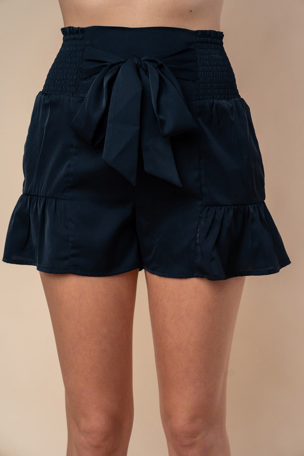 A woman is wearing the White Birch Full Size High Waisted Smocked Shorts, which feature a smocked elastic waistband with a large front bow and charming ruffle hem detailing.