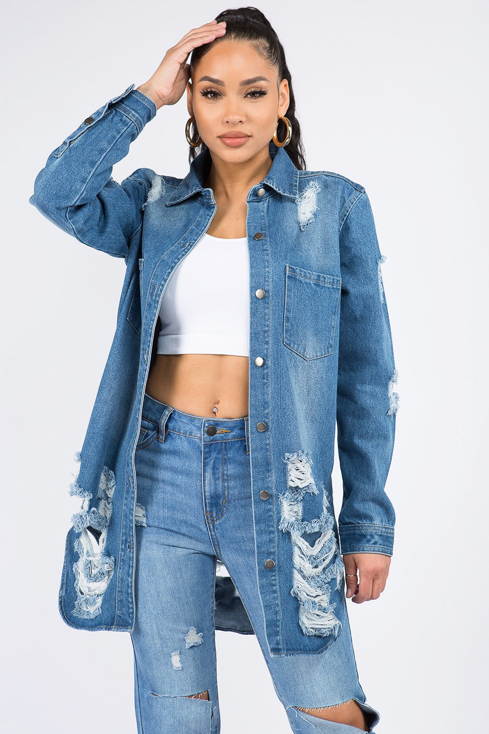 A woman wearing the American Bazi Distressed Button Down Denim Shirt Jacket and jeans paired with a white crop top exudes vintage-inspired charm as she poses with one hand on her head against a plain background.