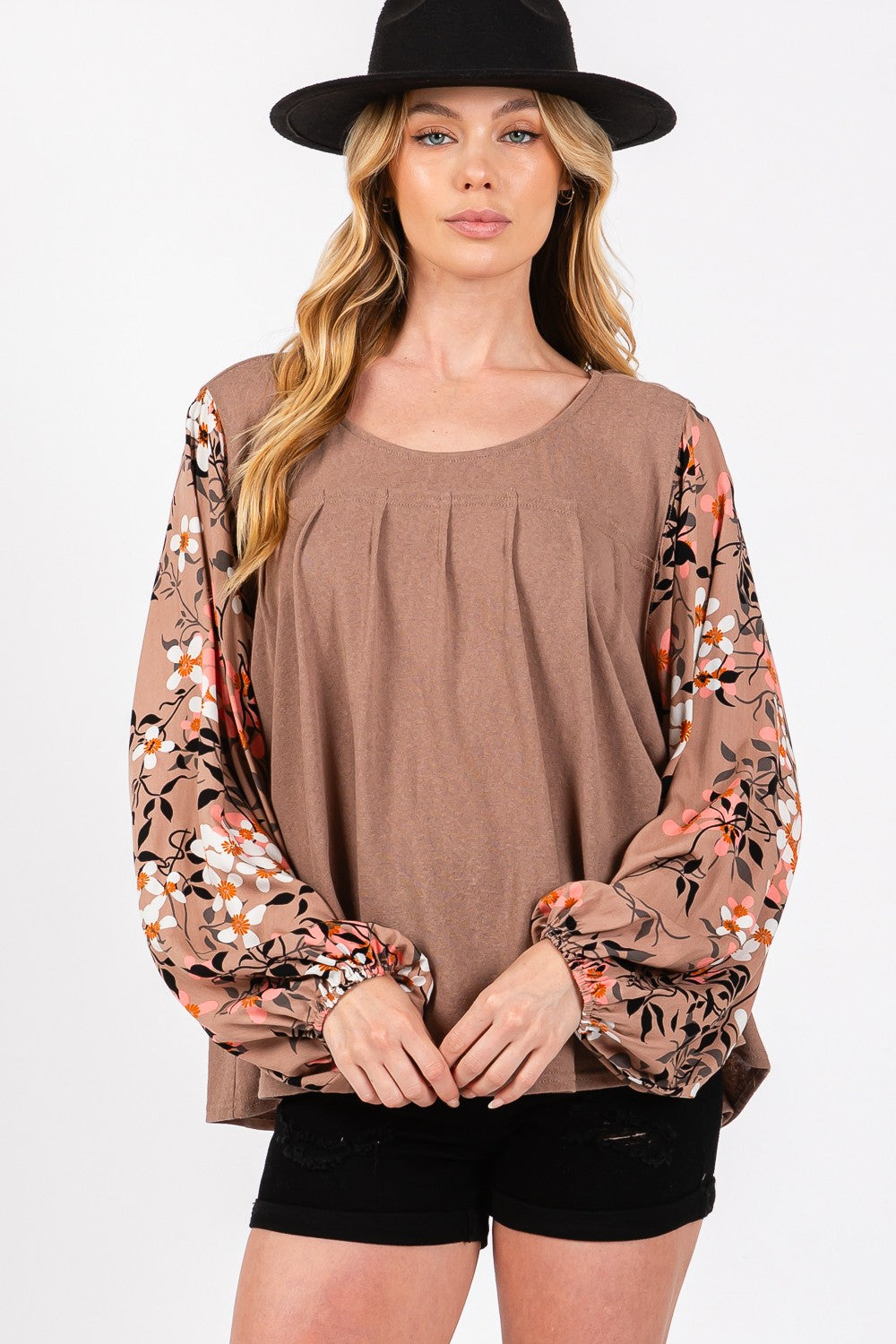 A person is wearing the SAGE + FIG Floral Long Sleeve Front Pleated Detail Blouse, paired with black shorts and a black hat, standing against a plain background.