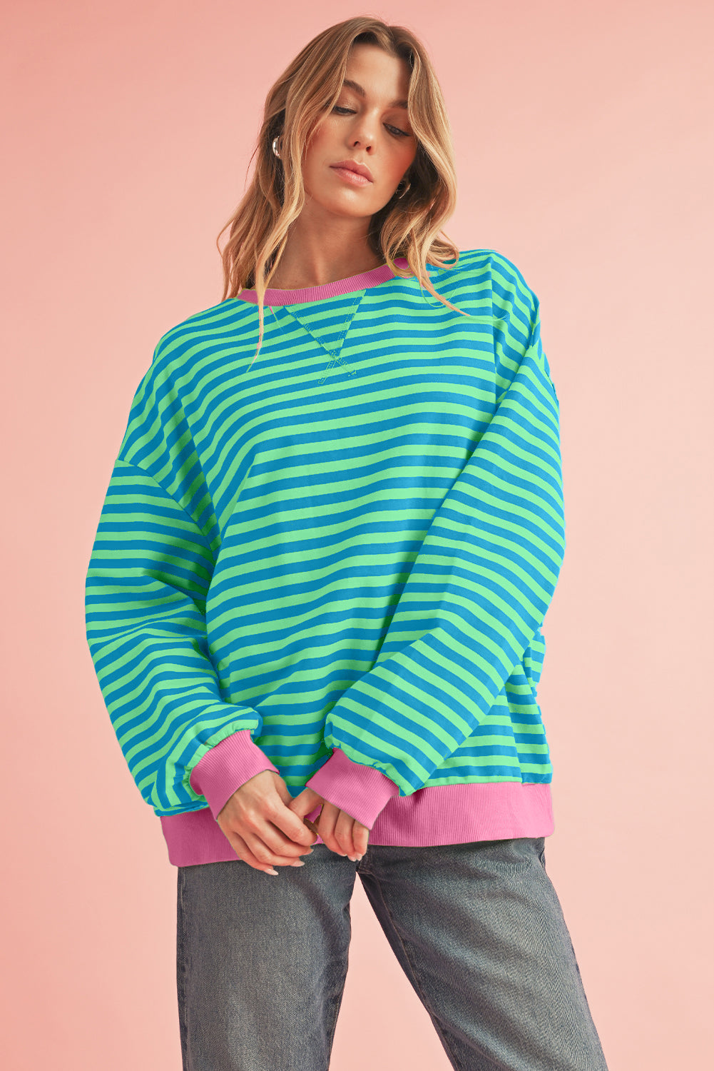 A person stands against a pink background wearing the Sky Blue Stripe Contrast Edge Oversized Pullover, featuring a loose, long-sleeved stripe design in green and sky blue hues with pink cuffs and neckline, paired with gray pants. Their hands are lightly clasped at their waist.