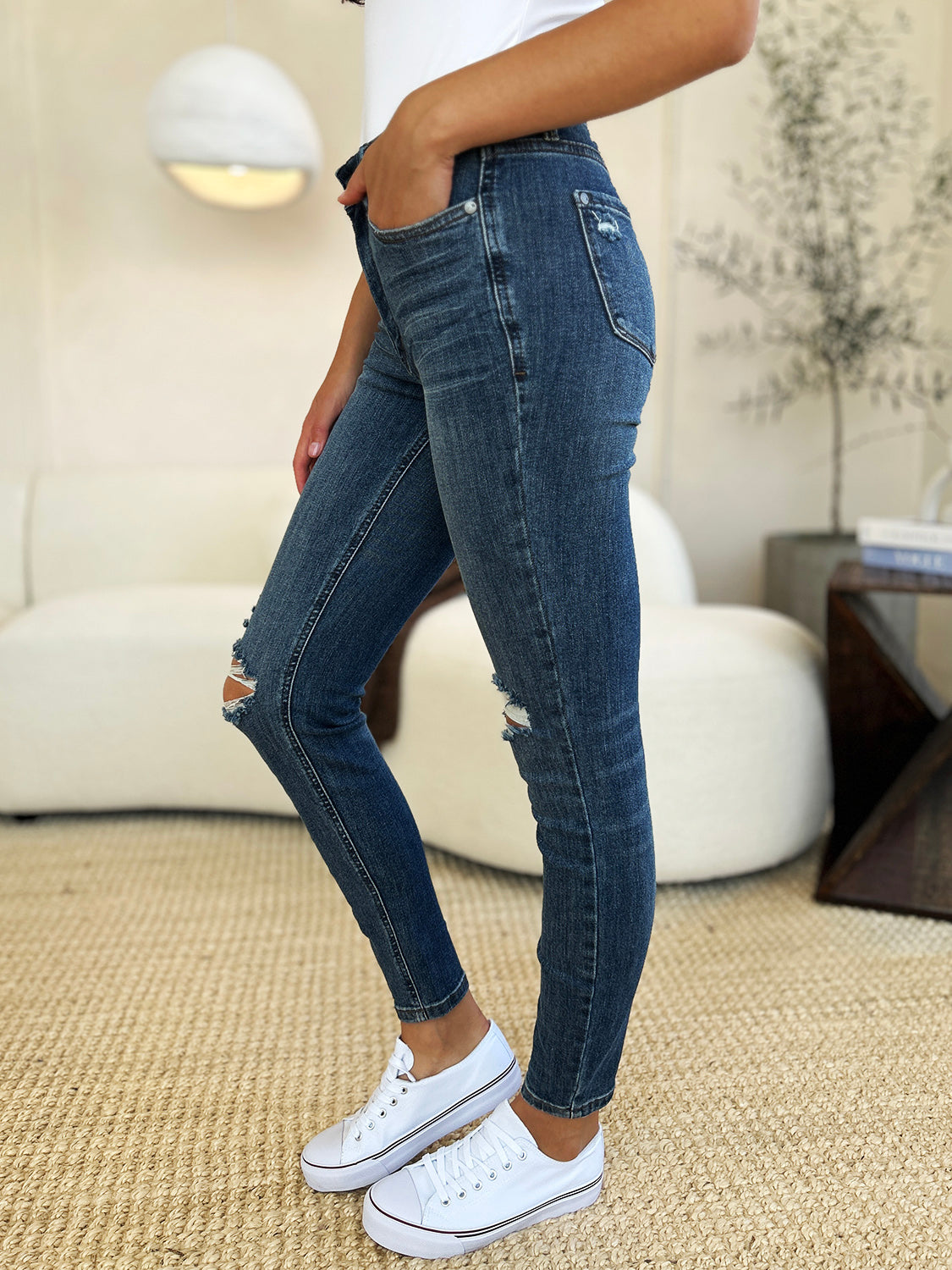 Person standing indoors wearing Judy Blue Full Size Mid Waist Distressed Slim Jeans, a black top, and black sneakers with white laces. There is a white sofa and a textured rug in the background.