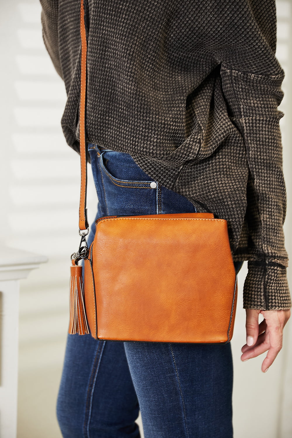 A person dressed in a dark sweater and blue jeans is holding the SHOMICO PU Leather Crossbody Bag with tassel detail, adding a touch of bohemian charm.